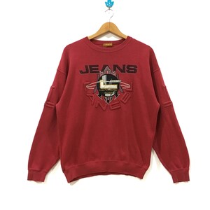 Könnte beinhalten: Ein rotes Sweatshirt mit dem Wort "JEANS" in Schwarz und einem grafischen Design in der Mitte. Das Sweatshirt hat lange Ärmel und einen Rundhalsausschnitt. Der Markenname "PANEE" ist ebenfalls sichtbar.