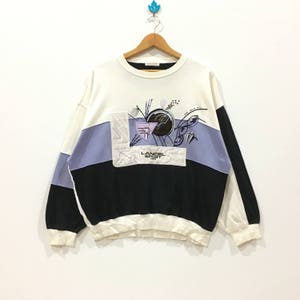 Könnte beinhalten: Ein Vintage-Sweatshirt mit Colorblock-Design. Der obere Teil ist weiß, mit einem lila und schwarzen Panel über der Brust. Das Sweatshirt hat eine abstrakte Grafik und den Text "LANCEL SPORT PARIS".