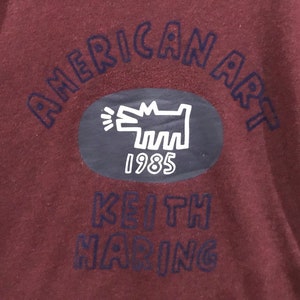 May include: Maroon fabric featuring the words "AMERICAN ART" in dark blue, arched across the top. An oval contains a white Keith Haring dog illustration and the year "1985." The words "KEITH HARING" are printed below.