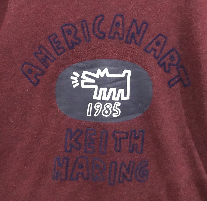 May include: Maroon fabric featuring the words "AMERICAN ART" in dark blue, arched across the top. An oval contains a white Keith Haring dog illustration and the year "1985." The words "KEITH HARING" are printed below.