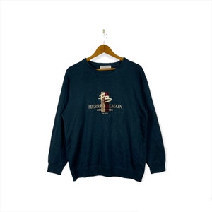 May include: A dark teal sweatshirt with the embroidered text "PIERRE BALMAIN COLLECTION PARIS" in gold and a stylized "PB" logo in red and brown. The sweatshirt is hanging on a wooden hanger.