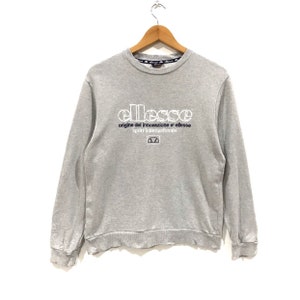 May include: A light gray sweatshirt with the brand name "ellesse" in white block letters across the chest. Below the brand name are the words "origine del innovazione e' ellesse sport internazionale" in white. The sweatshirt is hanging on a wooden hanger.