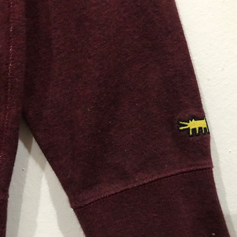 May include: Close-up of a maroon sweatshirt sleeve. The sleeve features a yellow embroidered logo of a stylized animal. The fabric has a textured appearance and is a solid maroon colour.