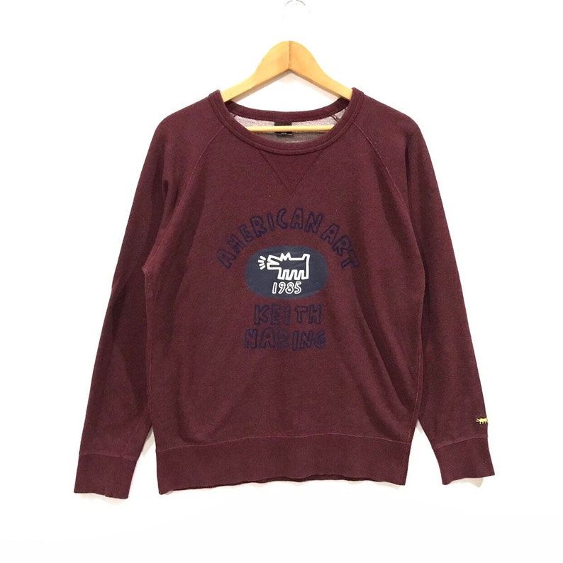 May include: A maroon sweatshirt with long sleeves, displaying the text "AMERICAN ART" above a blue oval with a white dog graphic and the year "1985". Below, the text reads "KEITH HARING". The sweatshirt is hanging on a wooden hanger.