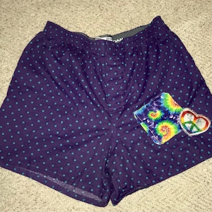 May include: Purple boxer shorts with a blue polka dot pattern. The shorts have a patch pocket on the right side with a tie-dye design and a peace sign heart.