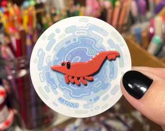 May include: A circular sticker featuring a bright orange shrimp illustration centred against a light blue and white swirling abstract background. The text ARTOULL is printed in small blue letters at the bottom left of the sticker design.