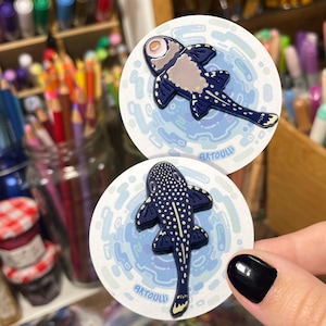 Puede incluir: Dos pegatinas redondas con un diseño de pez estilizado. El pez es azul oscuro con puntos blancos y detalles amarillos, sobre un fondo azul claro con un patrón de agua. El texto "ARTOULL" está impreso en la parte inferior de cada pegatina.