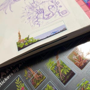 May include: A sketchbook with purple sketches of a flower and a cat-like face, and a sticker sheet featuring miniature terrarium designs with plants, trees, and a blue butterfly. The stickers are labeled "Stickers by Art".