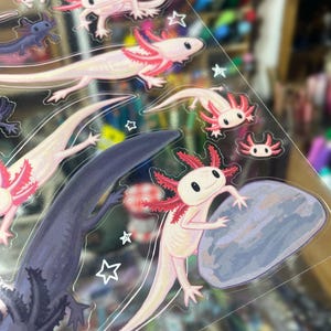 以下が含まれることがあります： ピンク、紫、白の色合いのアホロートルのイラストが描かれた透明なステッカーシート。岩を持っているアホロートルもいます。白い星の形がシート全体に散らばっています。アホロートルは赤いエラを持っています。