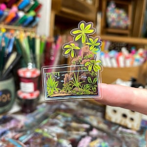 May include: A clear sticker featuring a miniature aquarium scene with green plants, rocks, and small fish. Above the aquarium is a cluster of green palm-like leaves. The sticker is held up by a finger.