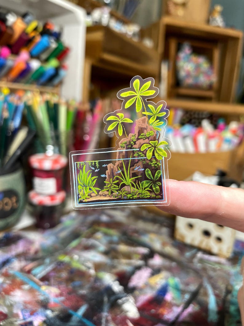 May include: A clear sticker featuring a miniature aquarium scene with green plants, rocks, and small fish. Above the aquarium is a cluster of green palm-like leaves. The sticker is held up by a finger.