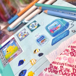 Peut inclure: Divers autocollants et articles de papeterie, dont des autocollants sur le th&egrave;me des poissons, un autocollant d'appareil portable r&eacute;tro et un autocollant de bouteille de savon. Des crayons de couleur et des stylos sont dispos&eacute;s autour des autocollants sur un cahier bleu clair.
