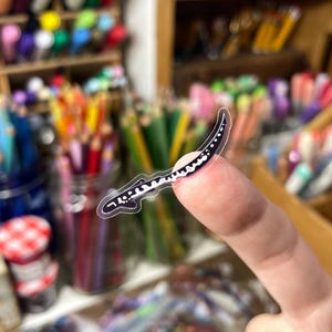 May include: A clear sticker featuring a black and white salamander design. The sticker is held up by a finger, with a blurred background of art supplies, including pencils and markers.