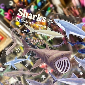 May include: Clear sticker sheet featuring various shark illustrations in shades of blue, gray, and brown. The sheet includes the text "Sharks" and "Stickers by Artoulli". The sharks are surrounded by white star accents.