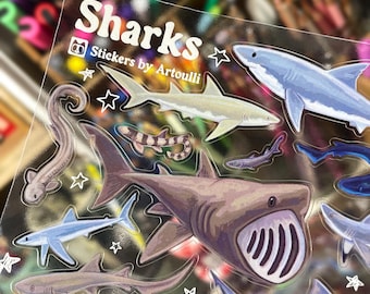 May include: A clear sticker sheet with illustrations of sharks in blue, grey, and brown. The sheet has the text "Sharks" and "Stickers by Artoulli". White stars surround the shark images.