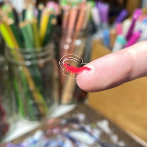 May include: A small, translucent sticker featuring a vibrant red shrimp illustration with a simple white outline. The sticker is tiny, shown resting on a fingertip, and displays a minimalist, hand-drawn aesthetic with a single black dot for an eye.