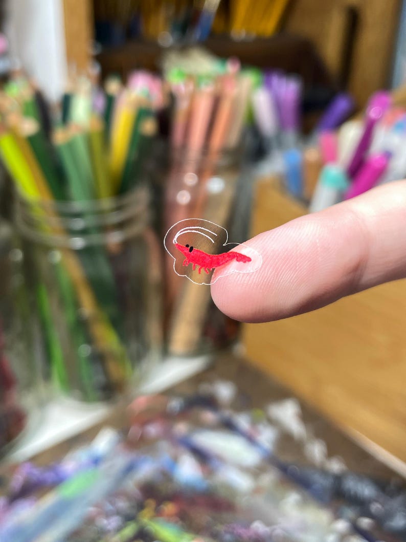 May include: A small, translucent sticker featuring a vibrant red shrimp illustration with a simple white outline. The sticker is tiny, shown resting on a fingertip, and displays a minimalist, hand-drawn aesthetic with a single black dot for an eye.