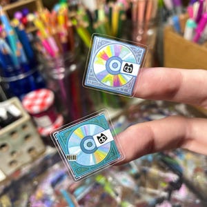 Peut inclure: Deux autocollants transparents repr&eacute;sentant un design de CD color&eacute; avec un petit logo noir et blanc. Les CD ont une palette de couleurs bleue et arc-en-ciel. Les autocollants sont tenus entre les doigts sur un fond flou de fournitures artistiques.