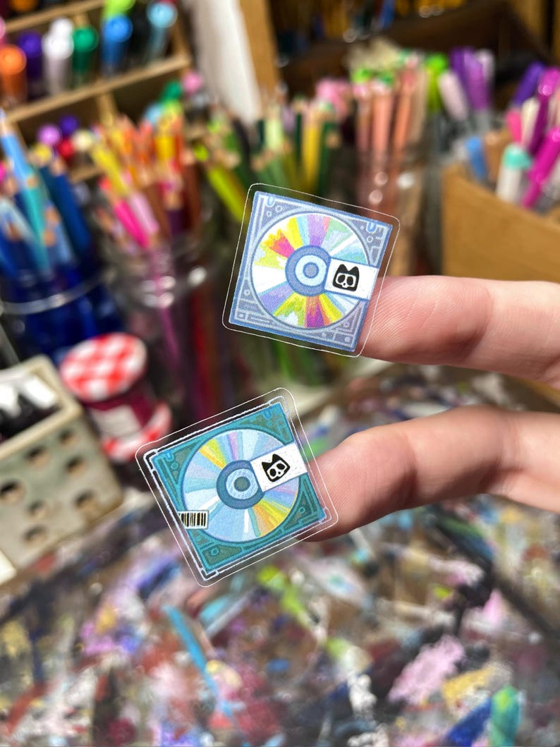Peut inclure: Deux autocollants transparents repr&eacute;sentant un design de CD color&eacute; avec un petit logo noir et blanc. Les CD ont une palette de couleurs bleue et arc-en-ciel. Les autocollants sont tenus entre les doigts sur un fond flou de fournitures artistiques.