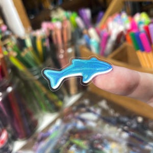 Peut inclure: Un autocollant de dauphin bleu avec un contour blanc est pr&eacute;sent&eacute; sur un fond flou de fournitures artistiques. L'autocollant est bleu clair avec des nuances bleues plus fonc&eacute;es, ce qui lui donne un effet 3D. L'autocollant est petit et semble &ecirc;tre en vinyle.