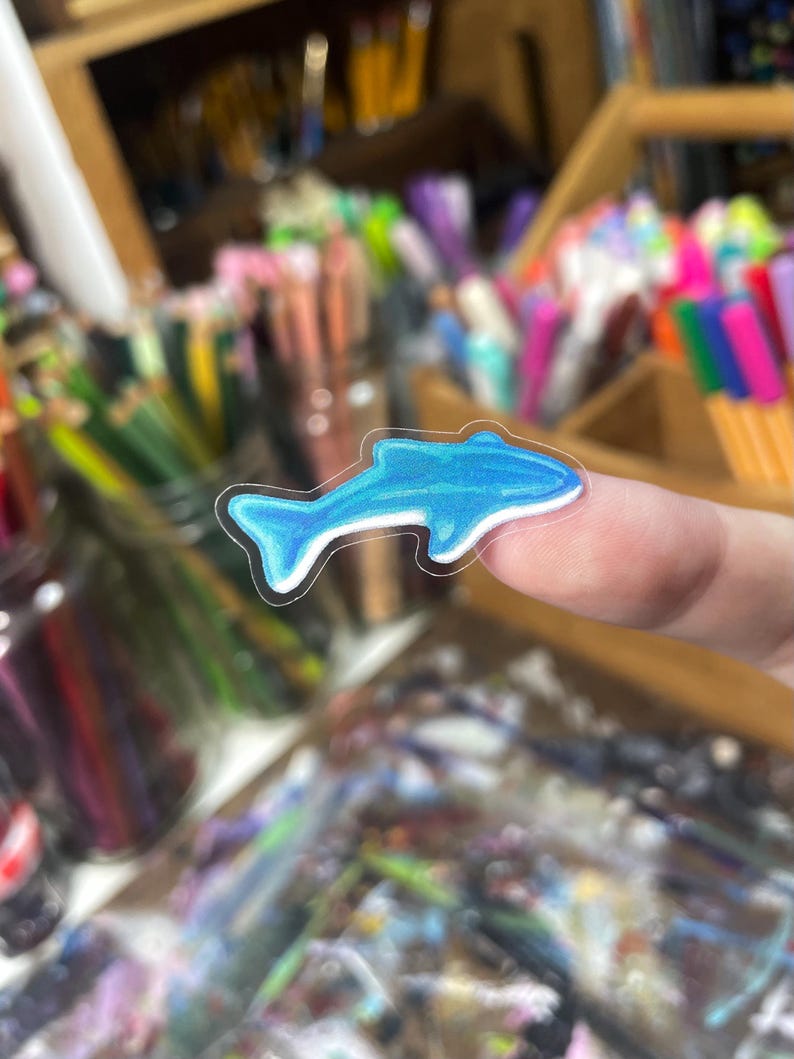 Peut inclure: Un autocollant de dauphin bleu avec un contour blanc est pr&eacute;sent&eacute; sur un fond flou de fournitures artistiques. L'autocollant est bleu clair avec des nuances bleues plus fonc&eacute;es, ce qui lui donne un effet 3D. L'autocollant est petit et semble &ecirc;tre en vinyle.