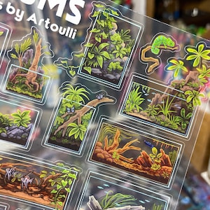 May include: Clear sticker sheet featuring miniature terrarium designs with vibrant green plants, a chameleon, and fish. The text "RIUMS by Artoulli" is visible at the top. Each terrarium is framed with a white border.