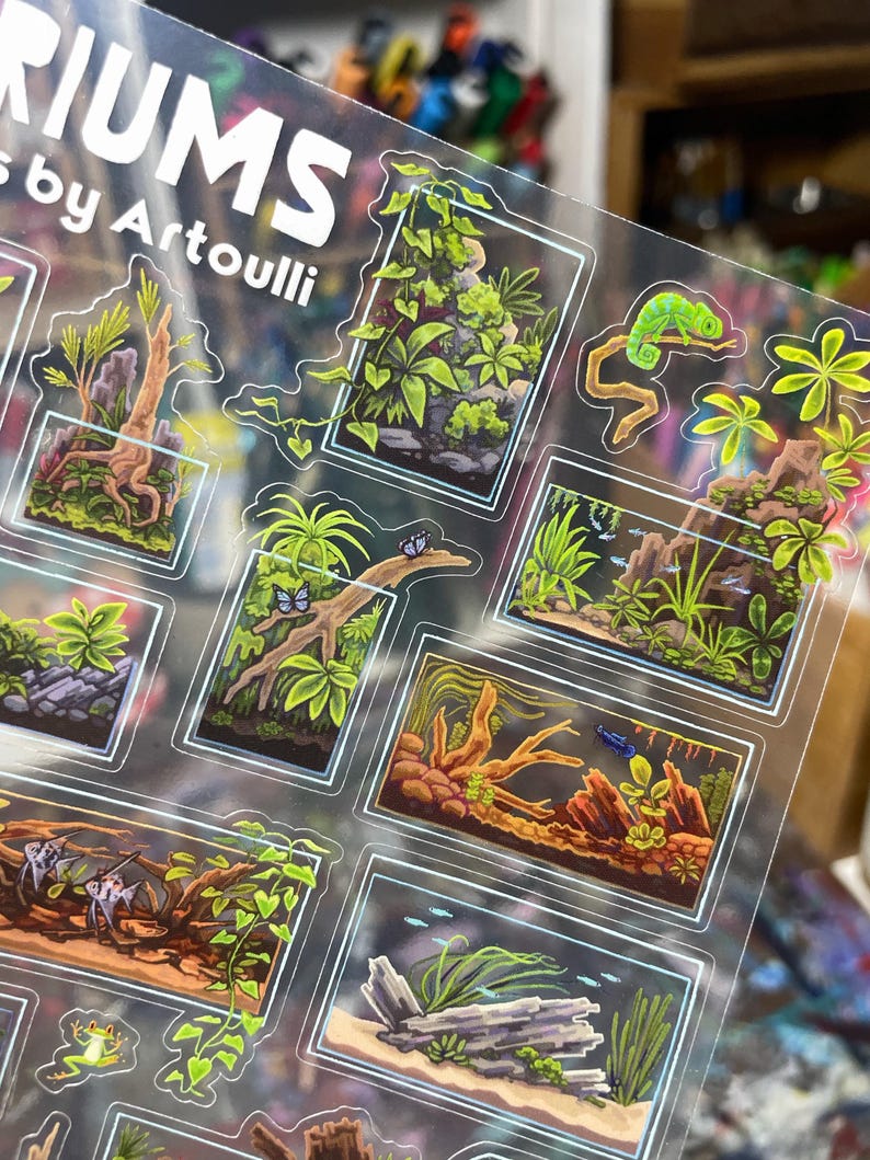 May include: Clear sticker sheet featuring miniature terrarium designs with vibrant green plants, a chameleon, and fish. The text "RIUMS by Artoulli" is visible at the top. Each terrarium is framed with a white border.