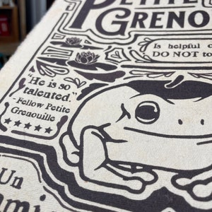 Może przedstawiać: Czarno-biała ilustracja żaby na kremowym tle. Ilustracja jest otoczona tekstem, kt&oacute;ry brzmi "Petite Grenouille" i "Jest tak utalentowany." Tekst zawiera r&oacute;wnież cytat od "Fellow Petite Grenouille" z pięcioma gwiazdkami.