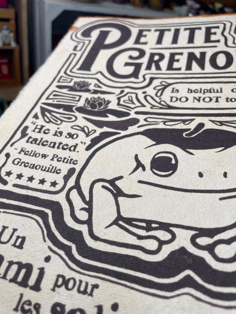 Może przedstawiać: Czarno-biała ilustracja żaby na kremowym tle. Ilustracja jest otoczona tekstem, kt&oacute;ry brzmi "Petite Grenouille" i "Jest tak utalentowany." Tekst zawiera r&oacute;wnież cytat od "Fellow Petite Grenouille" z pięcioma gwiazdkami.