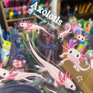 以下が含まれることがあります： ピンク、紫、白の色合いのアホロートルデザインが特徴の透明ステッカーシート。ステッカーはホログラフィック効果があり、「Axolotls Stickers by Artoulli」の文字が含まれています。
