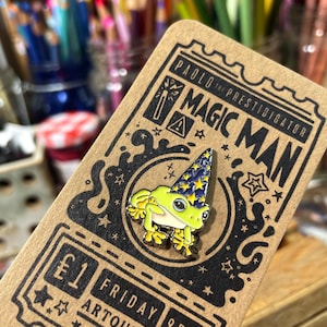 May include: A colorful enamel pin featuring a green frog wearing a blue wizard hat with yellow stars. The pin is on a brown card with the text "PAOLO the PRESTIDIGATOR MAGIC MAN". The card also has the text "&pound;1 FRIDAY 9PM ARTOULLI LLC".