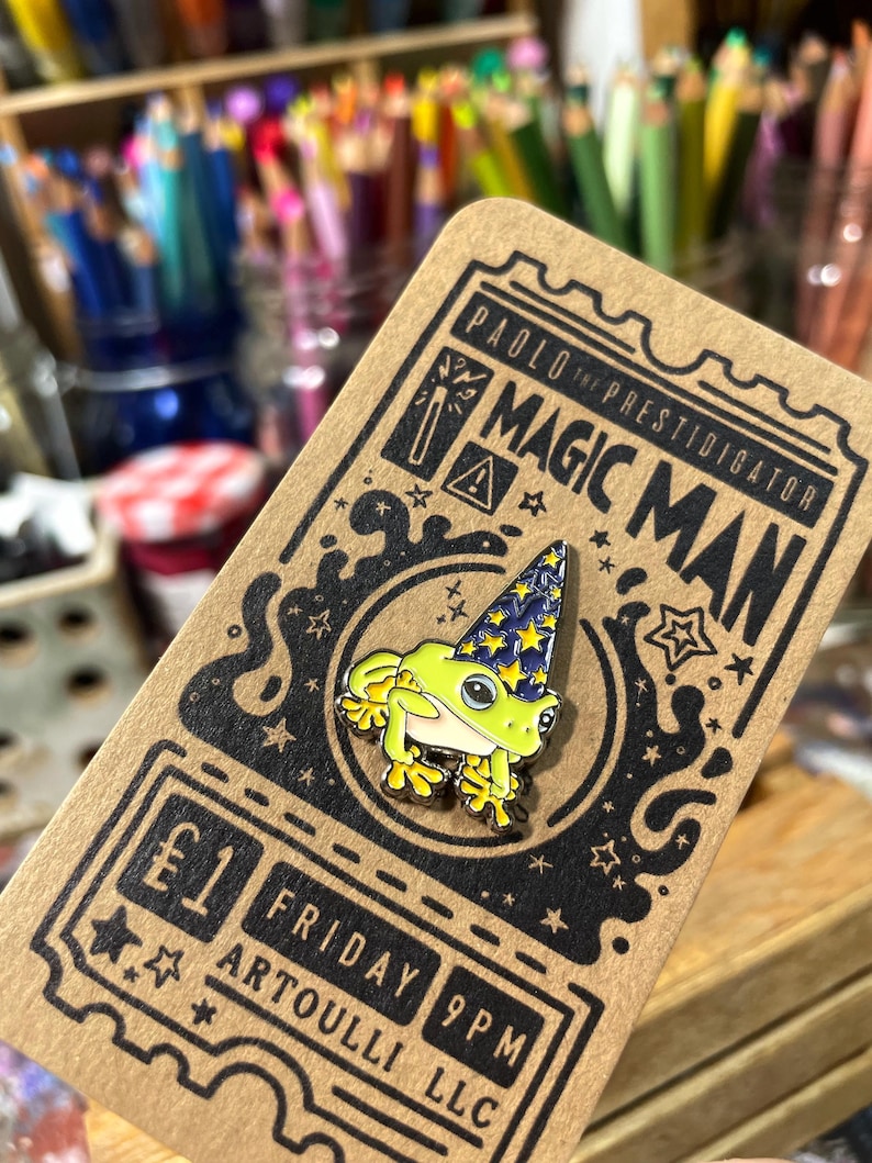 May include: A colorful enamel pin featuring a green frog wearing a blue wizard hat with yellow stars. The pin is on a brown card with the text "PAOLO the PRESTIDIGATOR MAGIC MAN". The card also has the text "&pound;1 FRIDAY 9PM ARTOULLI LLC".