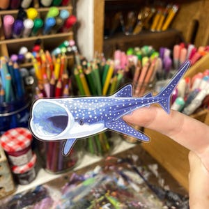 May include: A blue and white sticker of a whale shark with its mouth open. The sticker has a detailed illustration of the whale shark's spots and markings. The sticker is held up by a finger, with art supplies in the background.