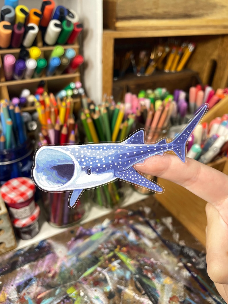 May include: A blue and white sticker of a whale shark with its mouth open. The sticker has a detailed illustration of the whale shark's spots and markings. The sticker is held up by a finger, with art supplies in the background.