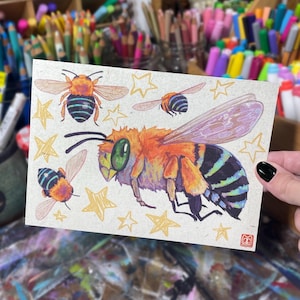 May include: A watercolor illustration of bees and stars on a beige card. The bees are orange, black, and green, with detailed wings. Several gold stars are scattered around the bees. The card is held by a hand with a black painted nail.