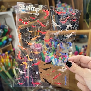 May include: A clear sheet of stickers featuring various shrimp illustrations in multiple colors and sizes. The top left corner displays the text Shrimp and Stickers by Artoulli. The shrimp are depicted in shades of red, orange, blue, black, and white.