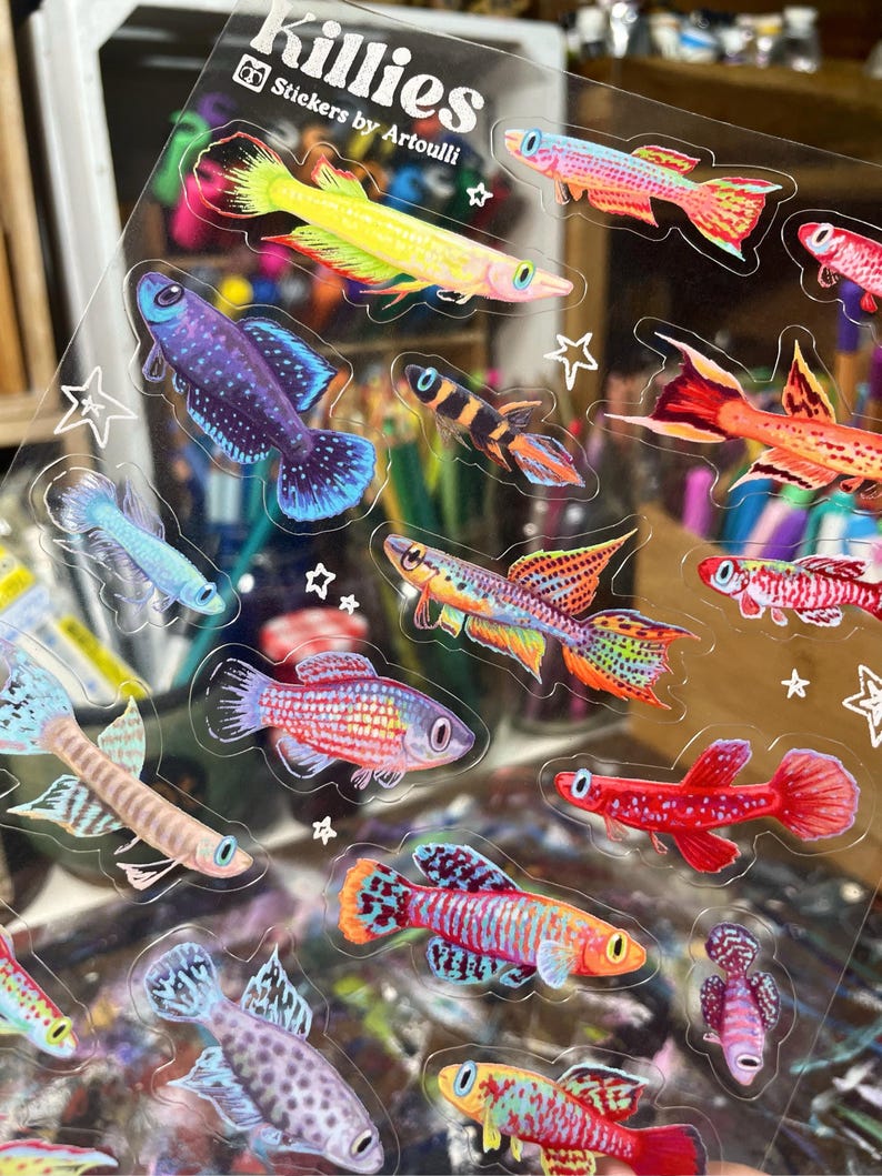 May include: A sheet of colorful fish stickers with the text "Killies Stickers by Artoulli". The stickers feature a variety of fish species in different colors and patterns, including red, orange, yellow, blue, green, and purple.