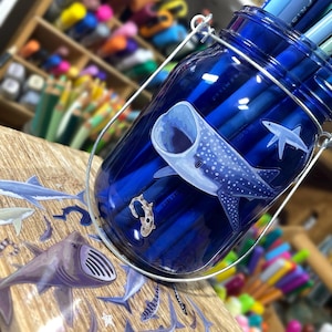 May include: A blue glass jar with a metal handle, filled with blue pencils. The jar is decorated with a whale shark and other sea creatures. The jar sits on a wooden surface.