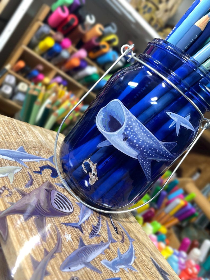 May include: A blue glass jar with a metal handle, filled with blue pencils. The jar is decorated with a whale shark and other sea creatures. The jar sits on a wooden surface.