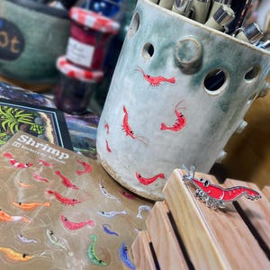 May include: A light green ceramic pencil holder with circular cutouts and small red shrimp illustrations, a sheet of shrimp stickers labeled Shrimp Stickers by Artoulli, and a single red shrimp enamel pin on a light wood block.