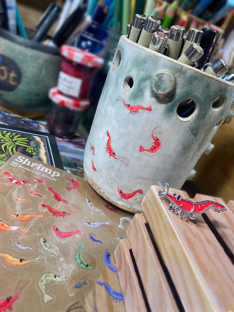 May include: A light green ceramic pencil holder with circular cutouts and small red shrimp illustrations, a sheet of shrimp stickers labeled Shrimp Stickers by Artoulli, and a single red shrimp enamel pin on a light wood block.