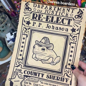 May include: A vintage-style poster with a cartoon frog wearing a sheriff's hat. The poster reads "Department of Justice, Re-Elect P.P. Johnson for County Sheriff". The poster has a distressed look and is printed on brown paper.