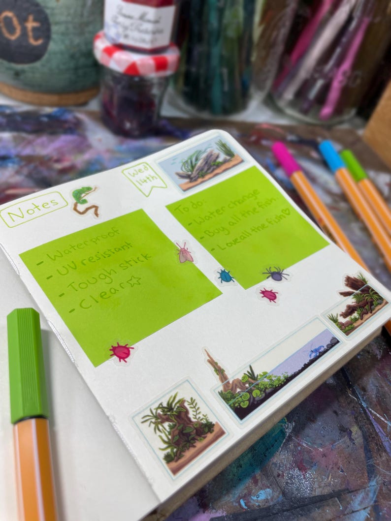 May include: Open notebook with green sticky notes and small stickers. The notes read "Waterproof, UV resistant, Tough stick, Clear." The "To do" list includes "Water change, Buy all the fish, Love all the fish." Colorful pens and jars are in the background.