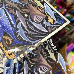 May include: Sheets of stickers featuring various shark illustrations in shades of blue, brown, and gray, set against a black background. The word "Sharks" is printed in white, with star accents. The stickers are designed by Artoulli.