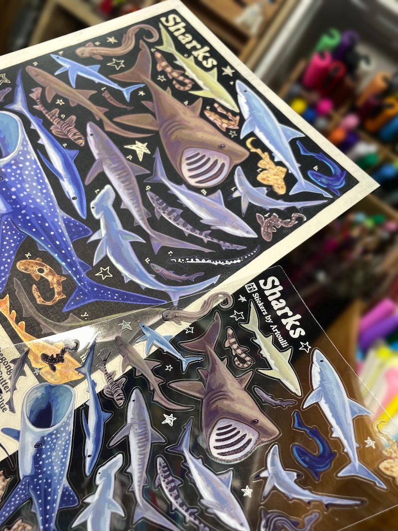 May include: Sheets of stickers featuring various shark illustrations in shades of blue, brown, and gray, set against a black background. The word "Sharks" is printed in white, with star accents. The stickers are designed by Artoulli.