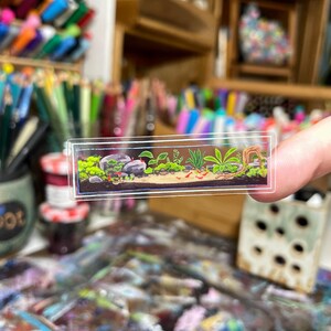May include: A miniature, rectangular aquarium sticker featuring a detailed underwater scene. The sticker showcases rocks, green plants, and small red creatures, all set against a clear background. The sticker is held up by a person's fingers.
