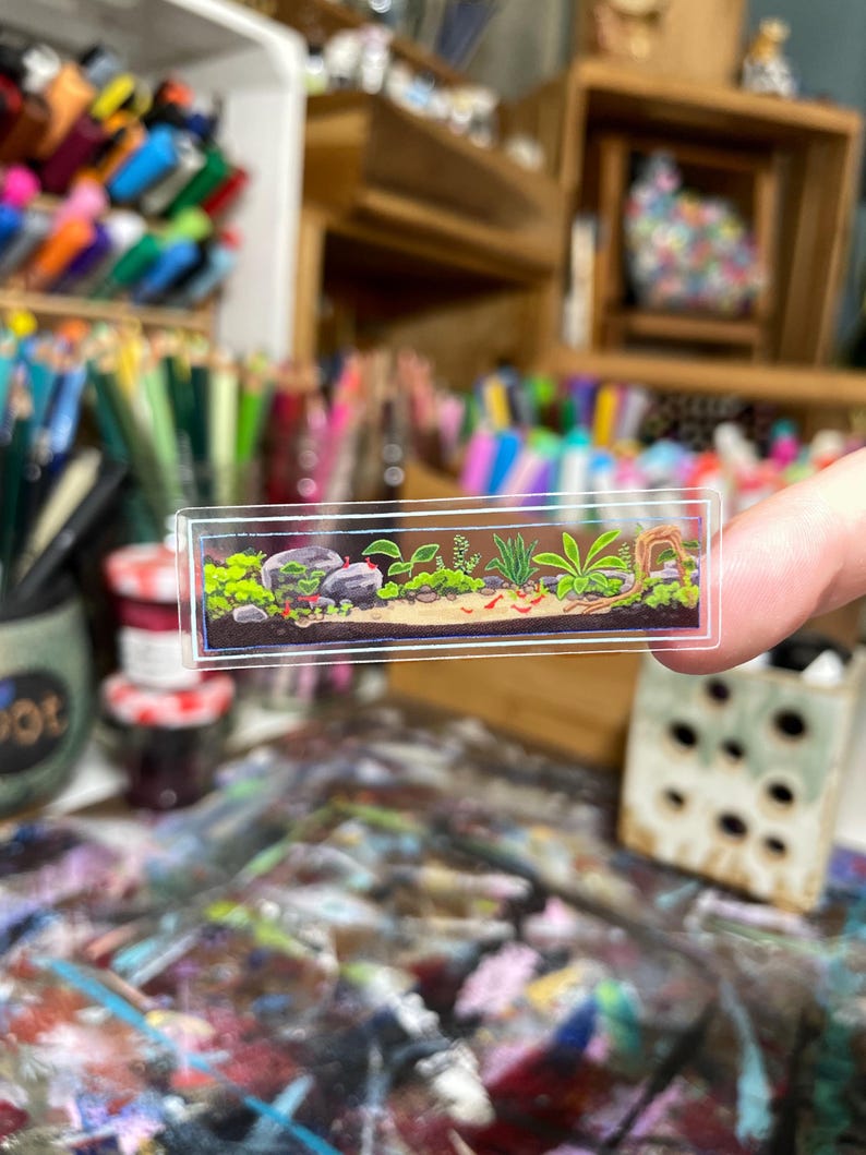 May include: A miniature, rectangular aquarium sticker featuring a detailed underwater scene. The sticker showcases rocks, green plants, and small red creatures, all set against a clear background. The sticker is held up by a person's fingers.