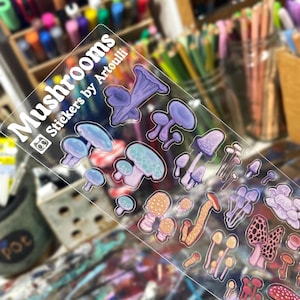 Peut inclure: Une planche d'autocollants transparents présentant diverses illustrations de champignons dans des tons de violet, bleu, orange et rose. Le texte "Mushrooms" et "Stickers by Artoulli" est imprimé en blanc sur le côté gauche.