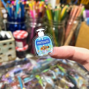 Peut inclure: Un autocollant transparent repr&eacute;sentant une bouteille bleue avec le mot "ARTOUIll" et un poisson-clown de dessin anim&eacute;. L'autocollant est tenu par un doigt, avec un arri&egrave;re-plan flou de fournitures d'art.