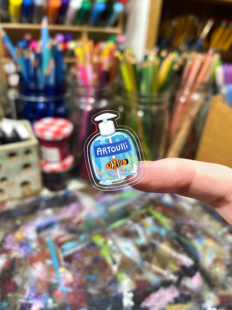 Peut inclure: Un autocollant transparent repr&eacute;sentant une bouteille bleue avec le mot "ARTOUIll" et un poisson-clown de dessin anim&eacute;. L'autocollant est tenu par un doigt, avec un arri&egrave;re-plan flou de fournitures d'art.